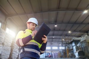A worker wearing a hard hat, safety glasses, and a high-visibility jacket writes on a clipboard inside a large industrial warehouse during engineering inspections NYC, with stacked materials and equipment visible in the background.
