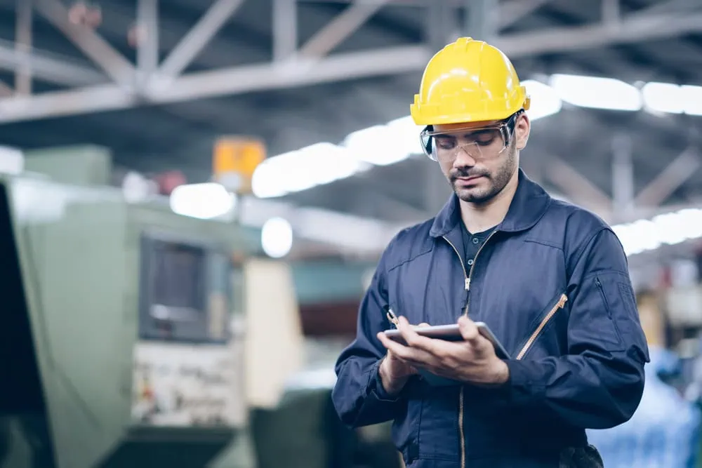 A male worker wearing a yellow hard hat, safety glasses, and a blue jumpsuit stands in a factory, using a digital tablet to conduct engineering inspections NYC, with machinery visible in the background.
