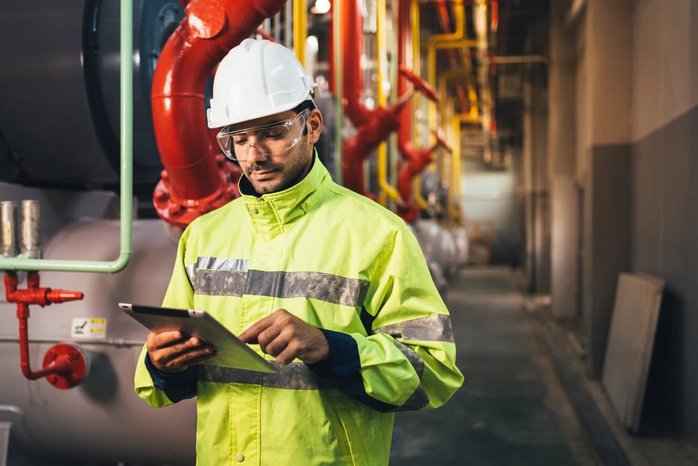 A worker in a high-visibility jacket, hard hat, and safety glasses uses a tablet to conduct engineering inspections NYC in an industrial setting with large pipes and machinery in the background.