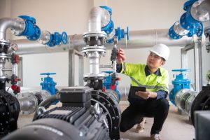 A worker wearing a hard hat and high-visibility jacket inspects and adjusts a valve on industrial pipes in a mechanical room, holding a tablet—demonstrating the precision of engineering inspections NYC professionals provide.