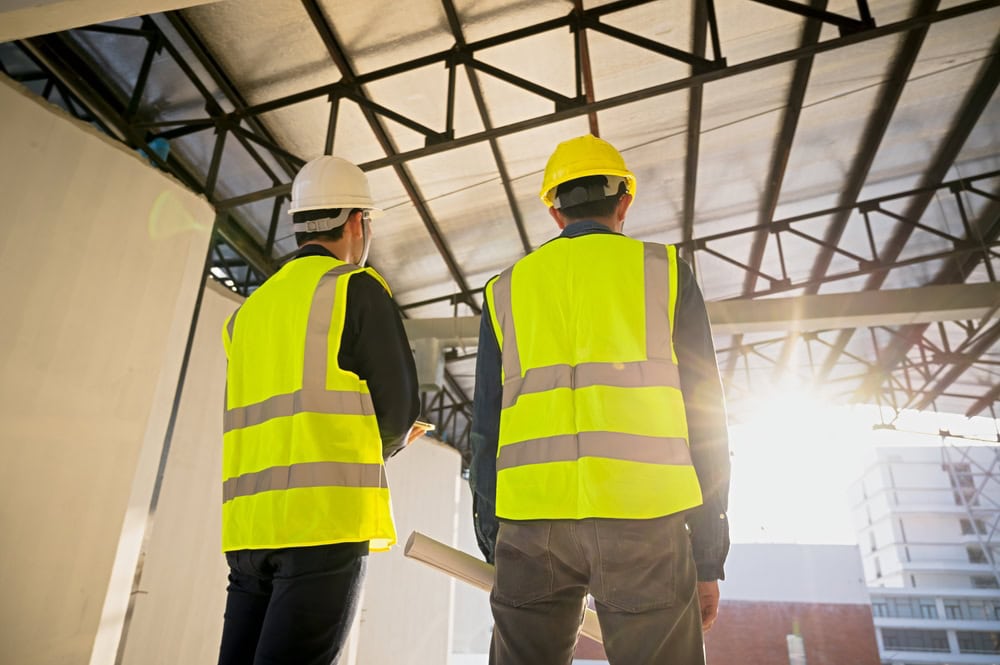 Two construction workers in yellow safety vests and hard hats stand inside a partially built structure, facing sunlight and discussing plans, as they prepare for engineering inspections NYC with exposed metal beams above them.