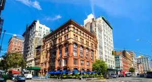 A vibrant city street corner with historic brick buildings, tall modern structures, traffic lights, and blue awnings over a sidewalk café—capturing the energy of NYC where engineering inspections NYC ensure safety under the clear blue sky.
