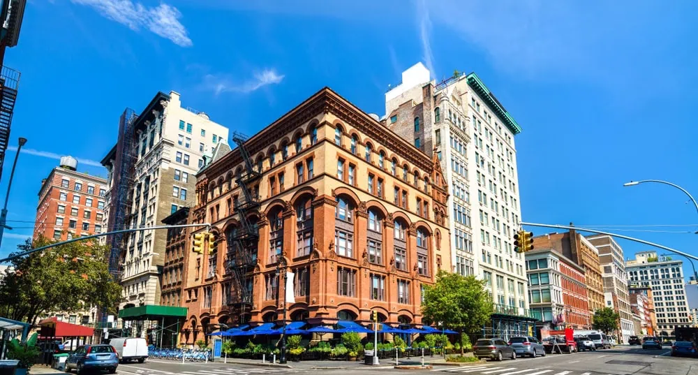 A vibrant city street corner with historic brick buildings, tall modern structures, traffic lights, and blue awnings over a sidewalk café—capturing the energy of NYC where engineering inspections NYC ensure safety under the clear blue sky.