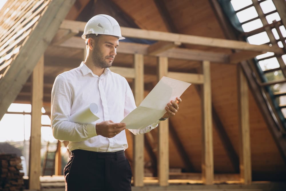A man wearing a white hard hat and shirt holds architectural plans while standing at a wooden building construction site, conducting engineering inspections NYC to ensure quality and progress.