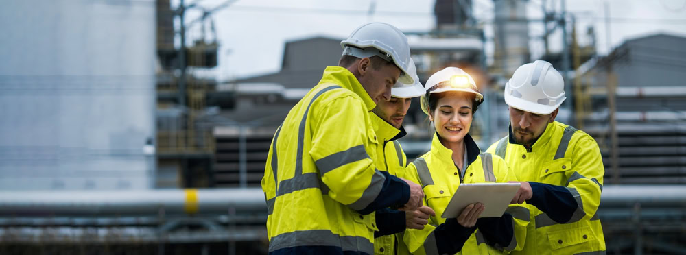 Four engineers wearing yellow safety jackets and white helmets stand outdoors at an industrial site, gathered around a tablet and smiling as they discuss engineering inspections NYC. Industrial equipment is blurred in the background.