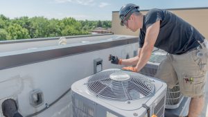 A technician in a hard hat and gloves conducts engineering inspections NYC, working on an air conditioning unit atop a rooftop, with tools clipped to his shorts and trees visible in the background.