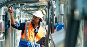 A worker in a hard hat, safety goggles, and orange vest uses a walkie-talkie and holds a tablet while performing engineering inspections NYC at an industrial facility.