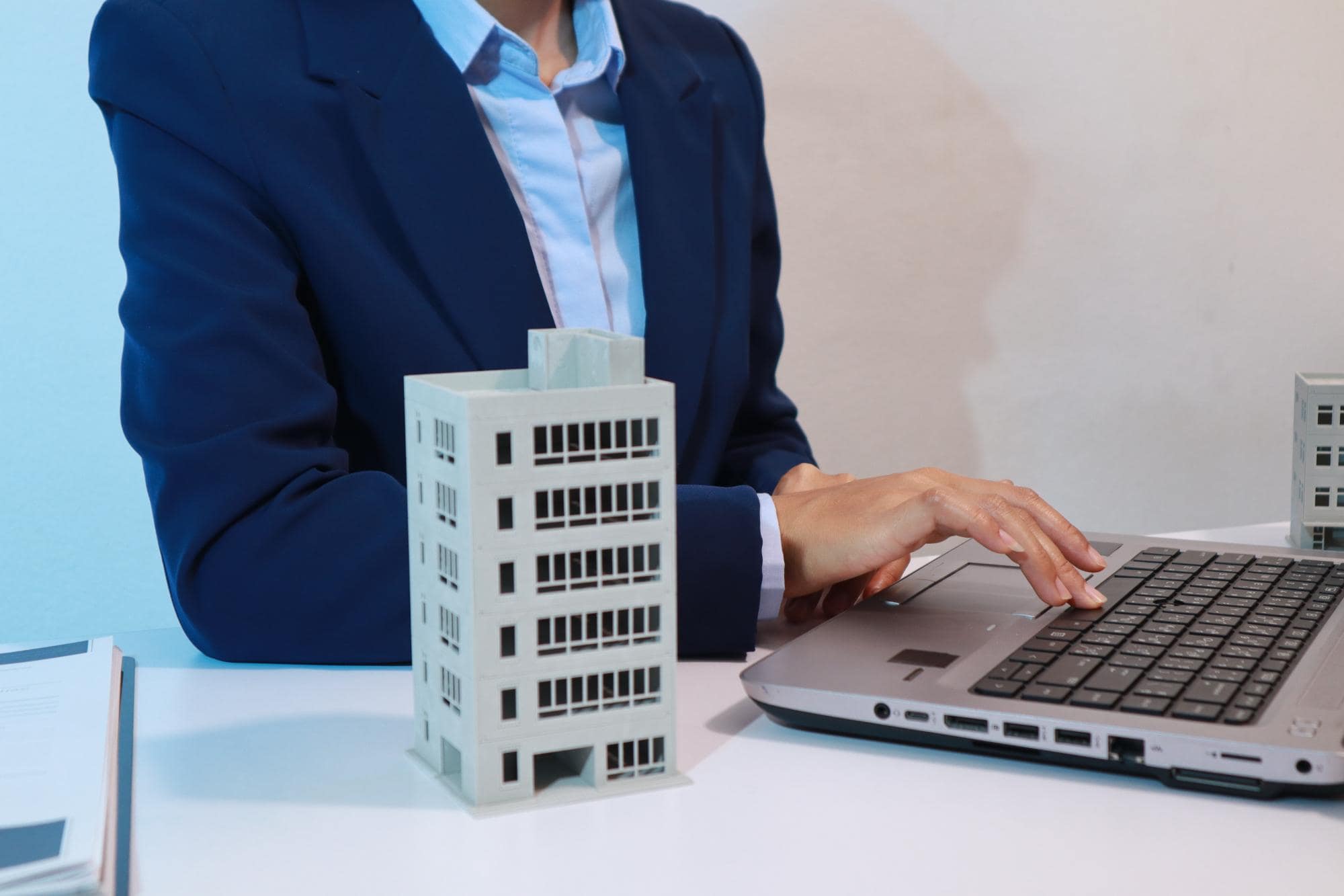A person in a blue suit sits at a desk using a laptop, with a small model of a modern apartment building beside them—illustrating the detail and professionalism required for engineering inspections NYC.