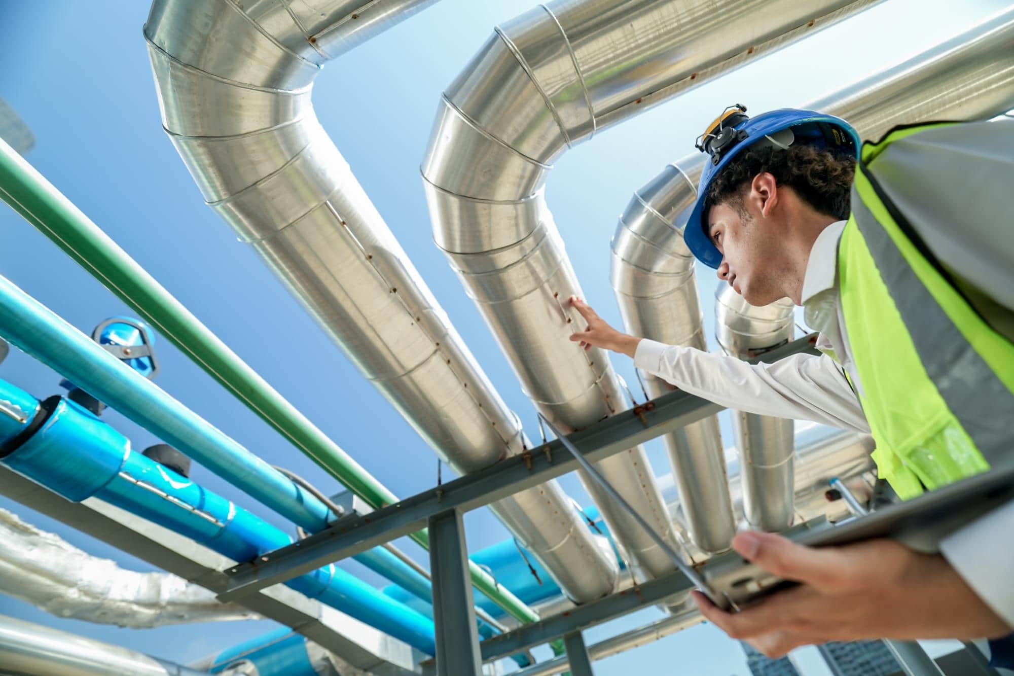 A worker in a hard hat and safety vest inspects large metal pipes outdoors under a blue sky, holding a tablet and reaching up toward the pipes—demonstrating expert engineering inspections NYC relies on.