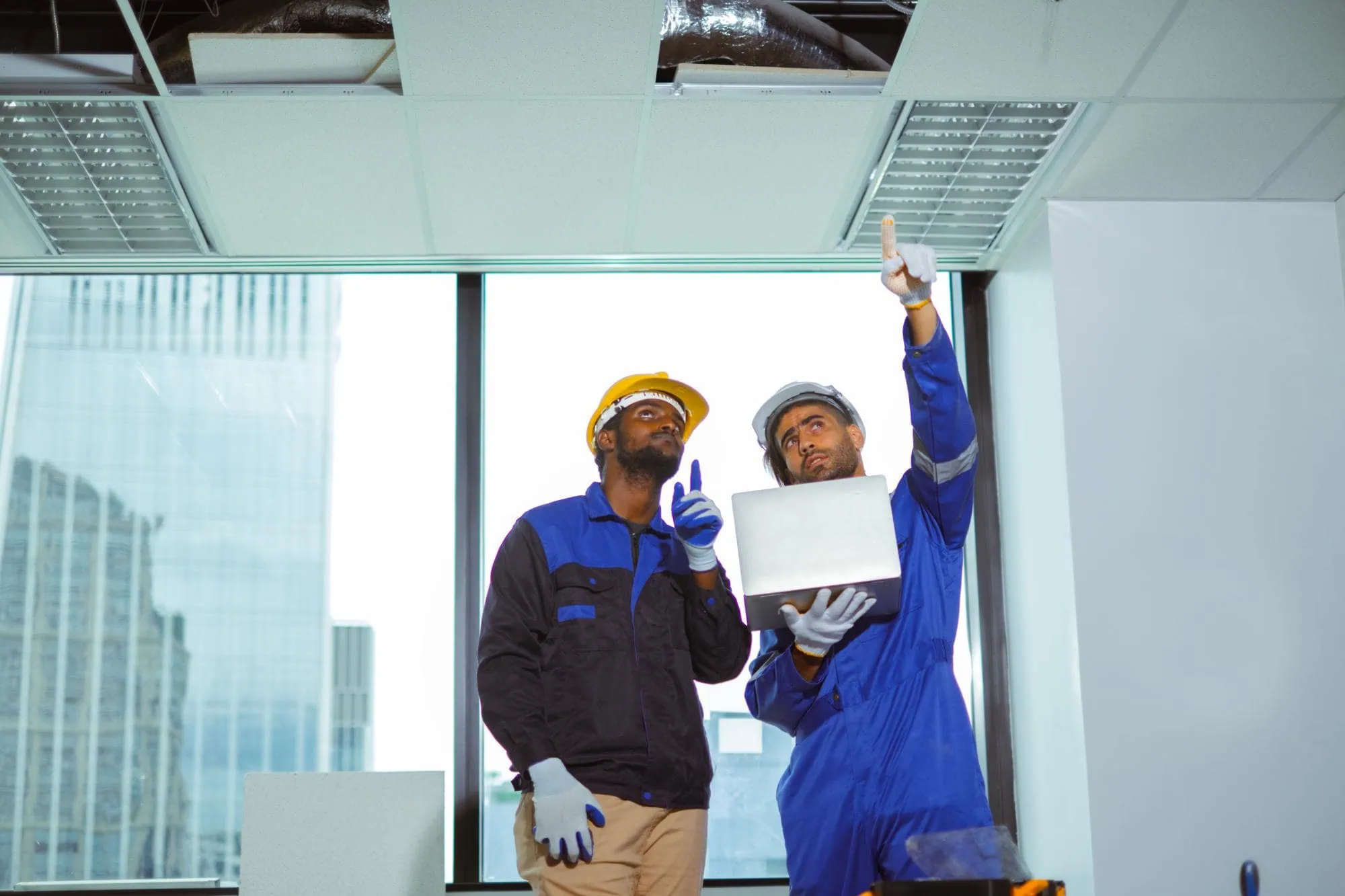 Two workers in protective gear conduct engineering inspections NYC, examining an office ceiling with a laptop and pointing upward. Large windows reveal tall buildings outside, highlighting professional inspection services NYC in action.