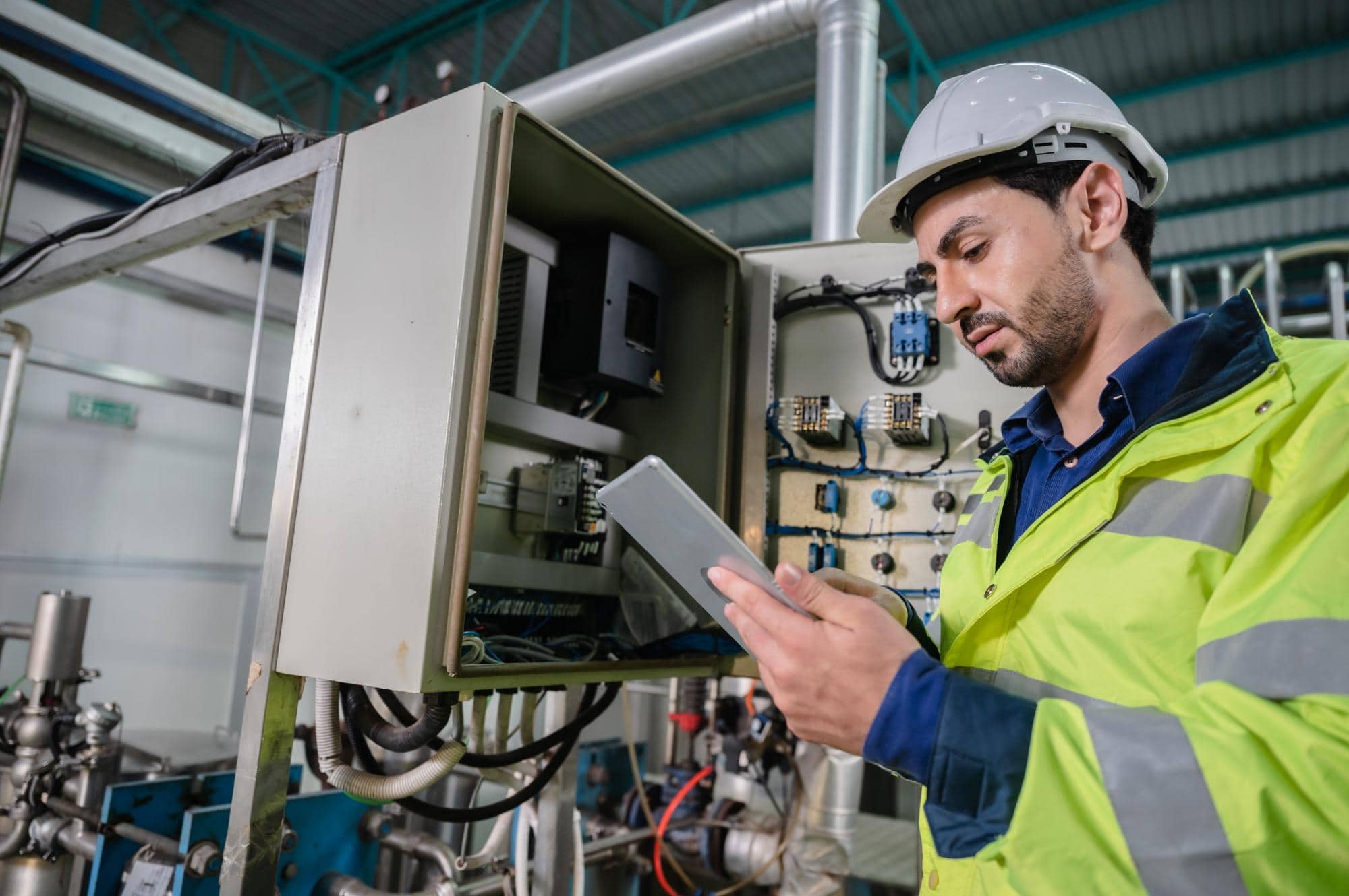 A male engineer in a high-visibility jacket and helmet uses a tablet while performing engineering inspections NYC on electrical equipment in an industrial facility.