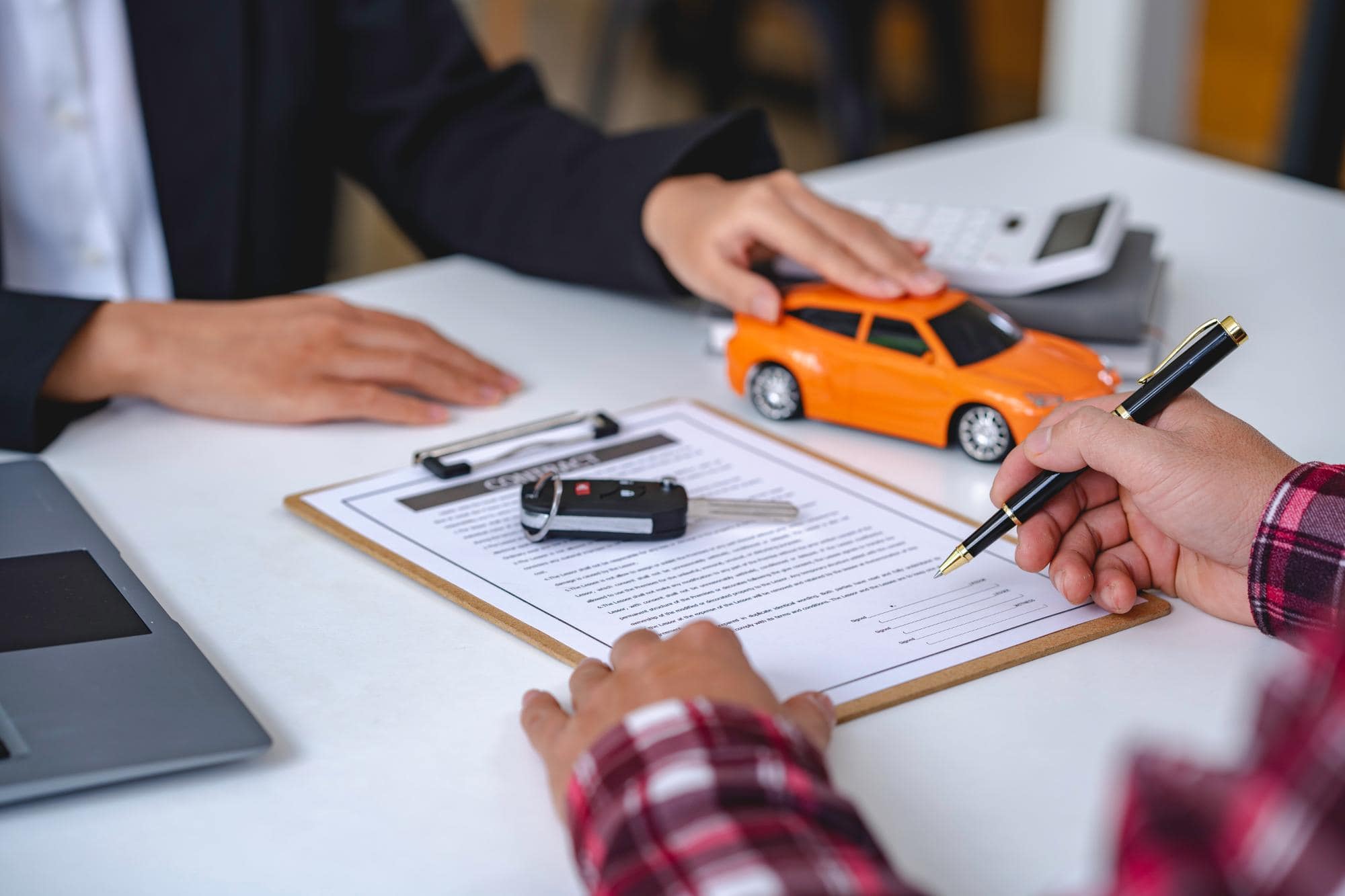 A person signs a car purchase or lease agreement on a clipboard, with car keys, a toy car, and a calculator nearby—while another in business attire sits across, highlighting the importance of inspection services NYC in every transaction.