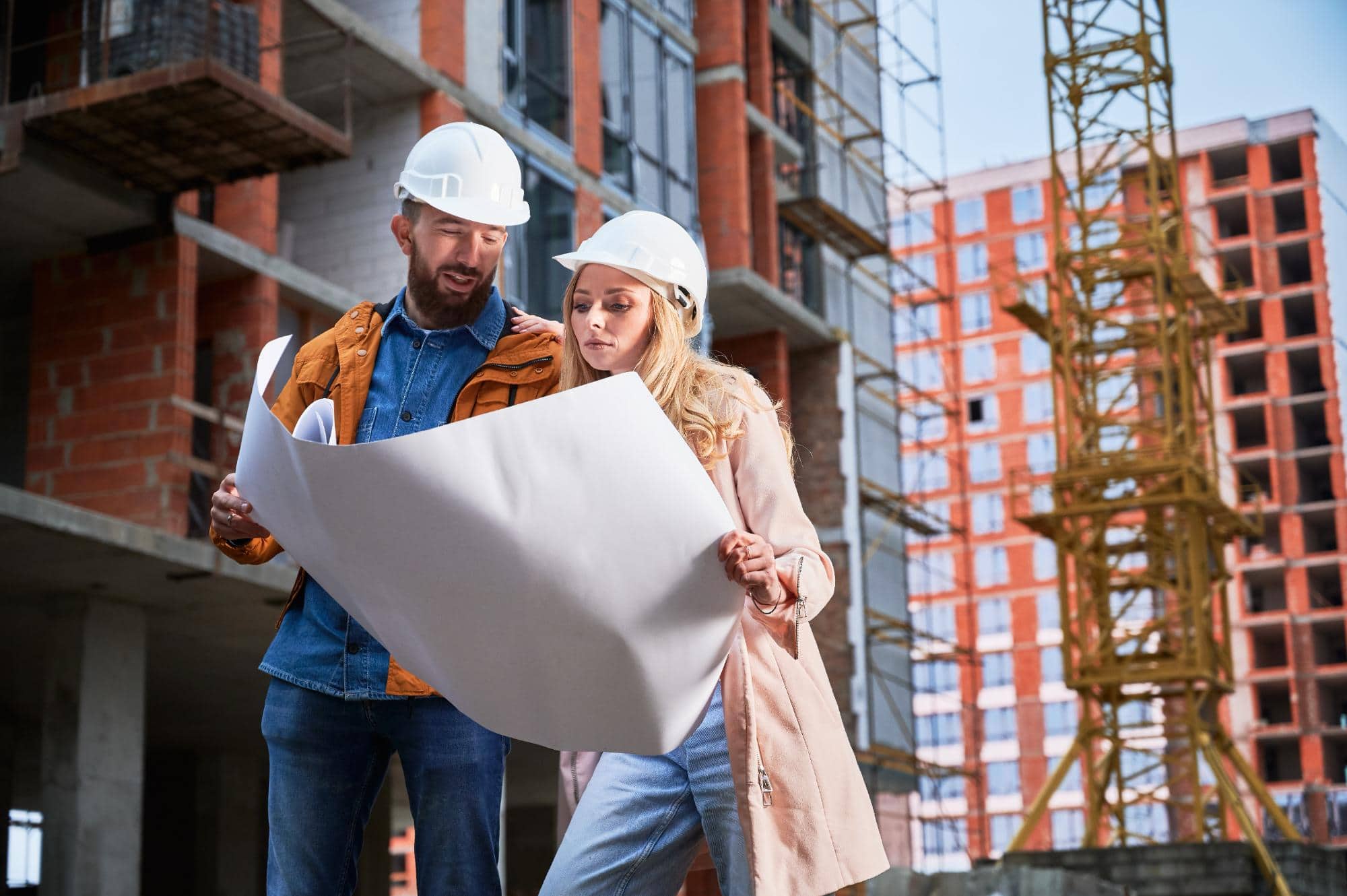 Two construction workers wearing hard hats examine architectural blueprints at a building site, where a crane and partially constructed brick buildings hint at ongoing engineering inspections NYC to ensure quality and safety.