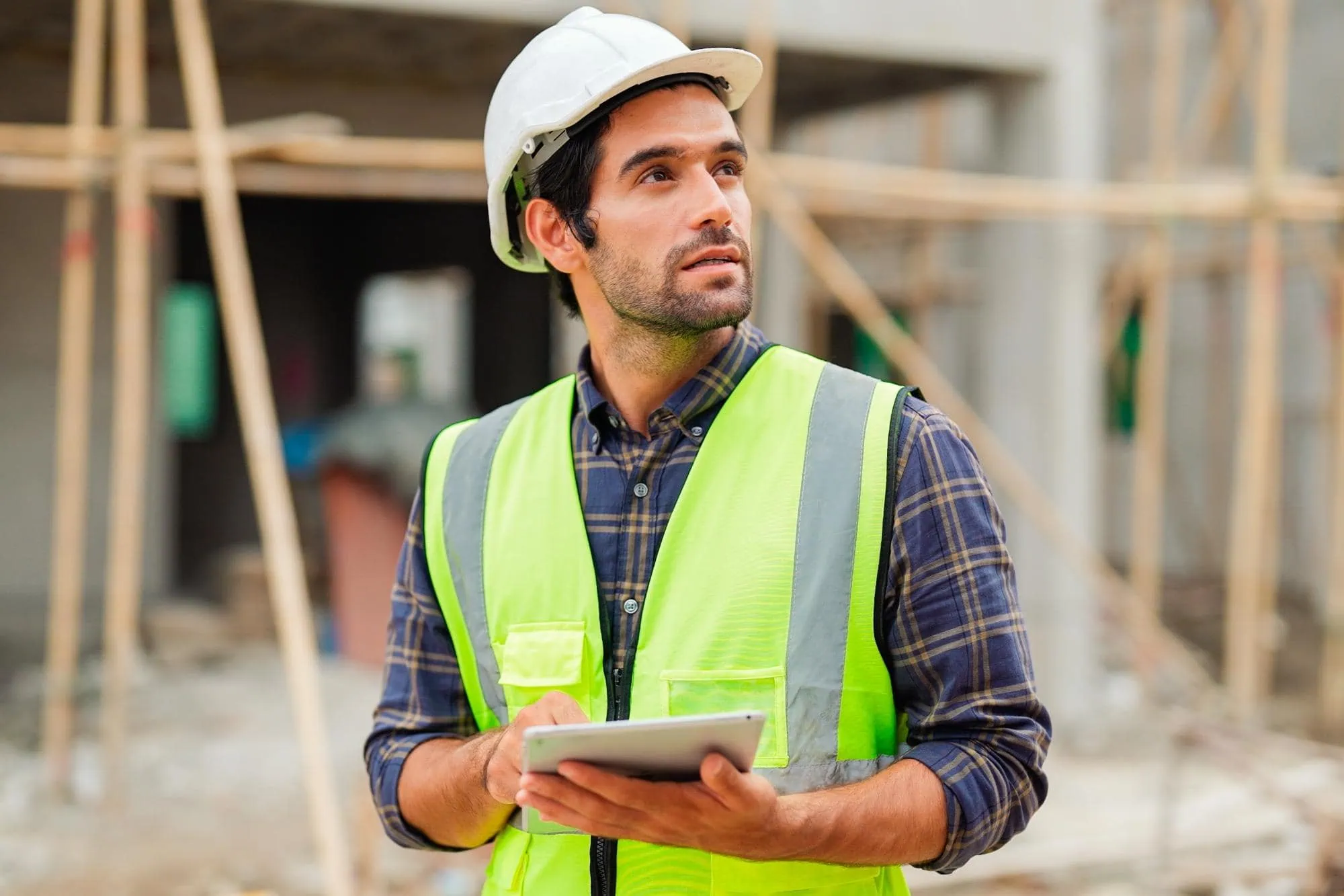 A construction worker in a white hard hat and neon safety vest holds a tablet and looks upward, standing at a construction site with scaffolding—ideal for engineering inspections NYC.