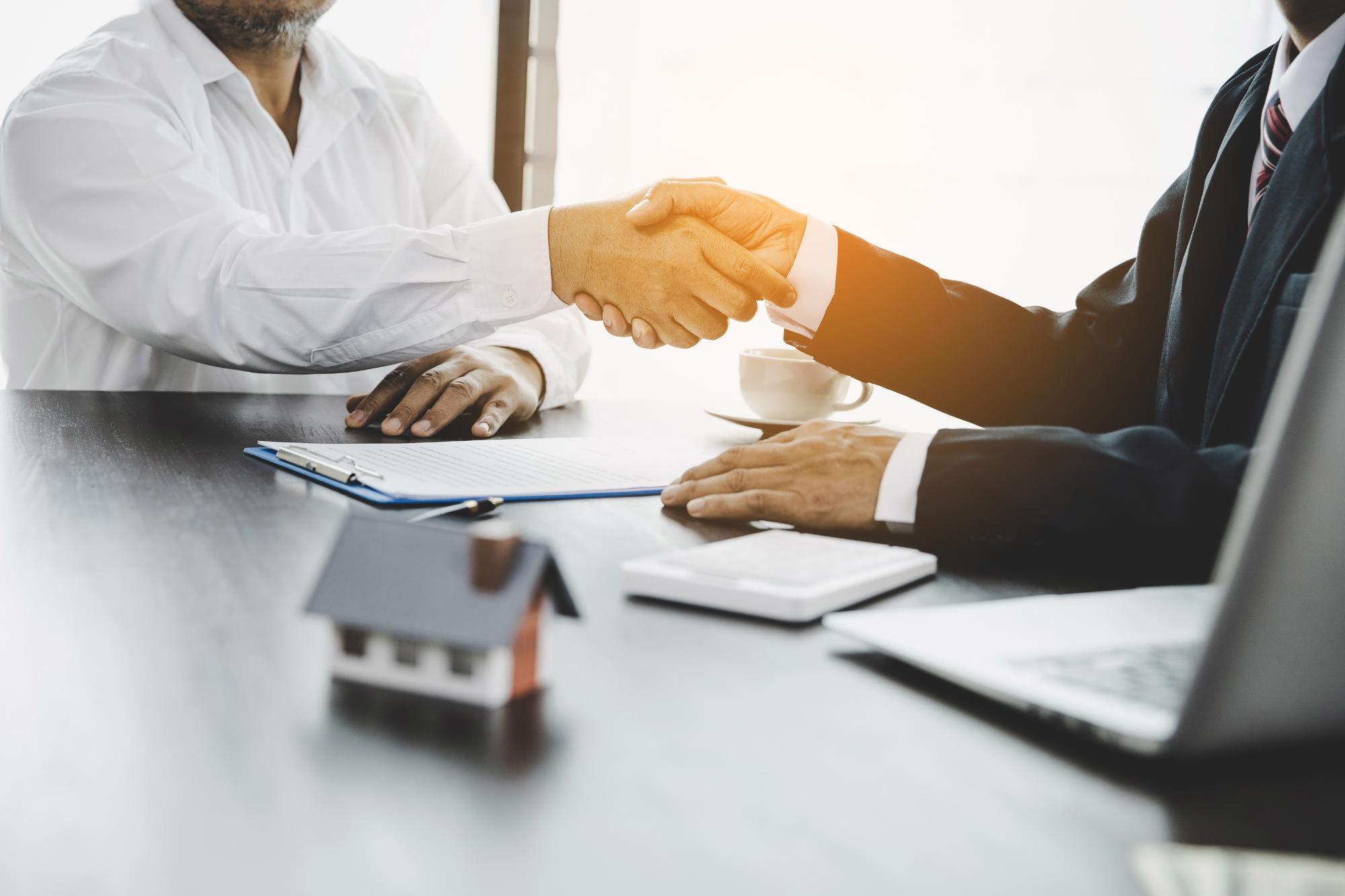 Two people in business attire shake hands across a desk with contract papers, a coffee cup, a small model house, and a laptop, suggesting an engineering inspections NYC or real estate agreement.
