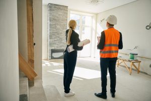 Two people, one in a suit holding blueprints and the other in a hard hat and orange safety vest, stand in a partially renovated room with large windows, discussing progress during engineering inspections NYC.