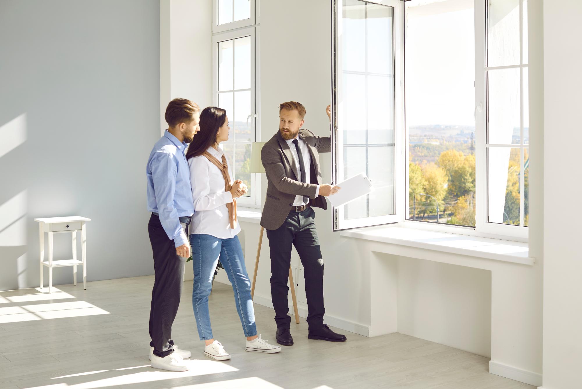 A real estate agent shows a young couple the features of a bright, modern apartment with large windows and lots of natural light, highlighting recent engineering inspections NYC as they listen attentively by the open window.